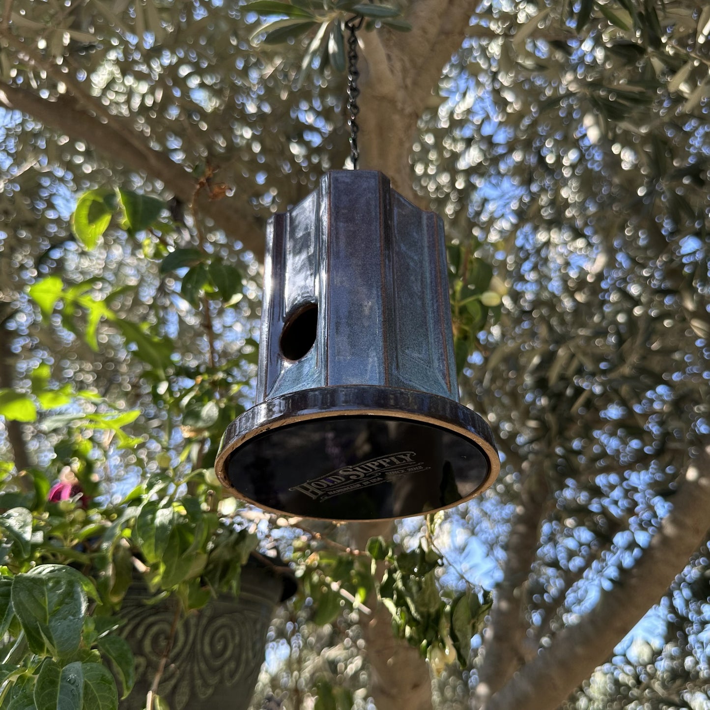 A hanging metal birdhouse with a weathered blue finish, suspended by a black chain in an outdoor garden setting with trees and plants in the background.