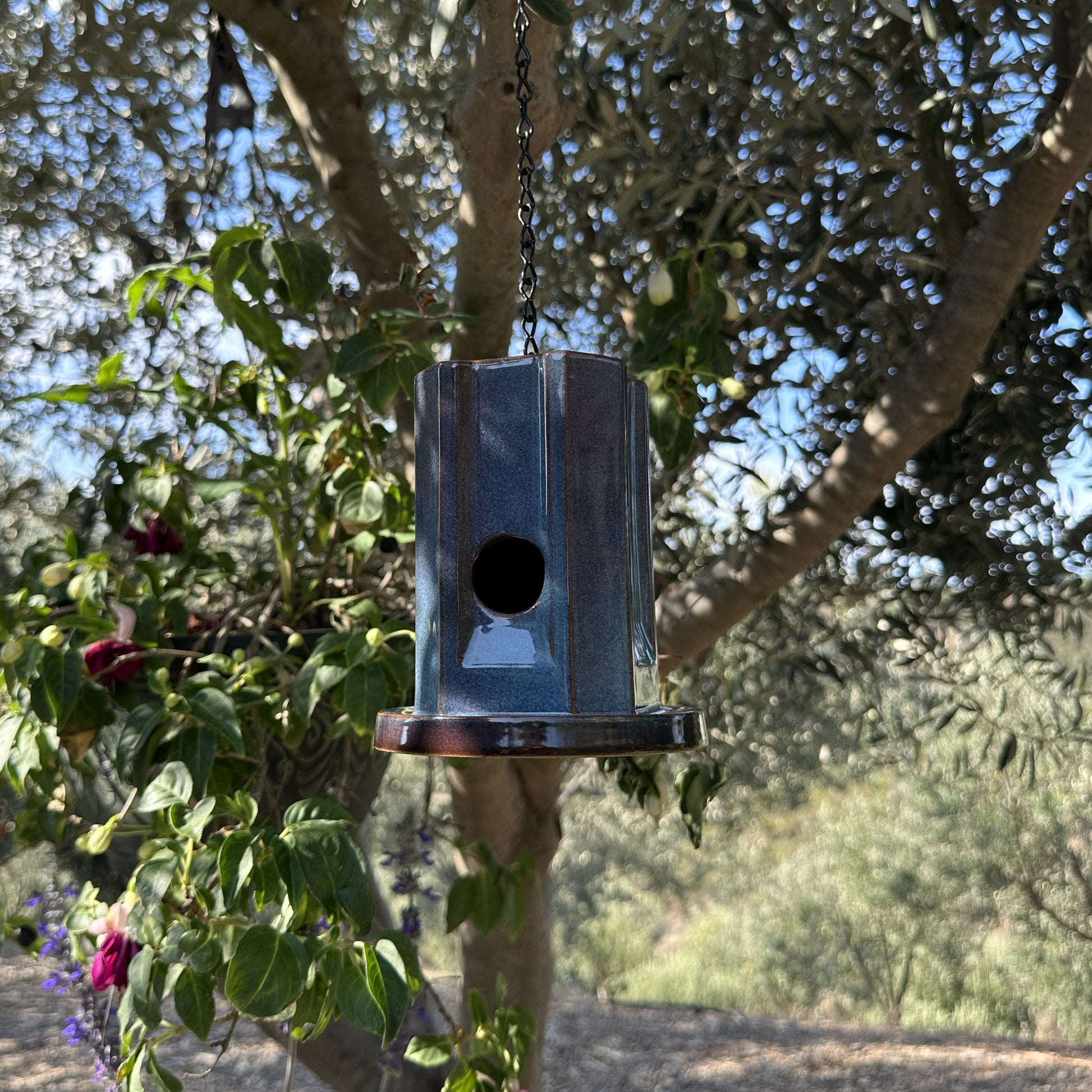 A hanging metal birdhouse with a weathered blue finish, suspended by a black chain in an outdoor garden setting with trees and plants in the background.