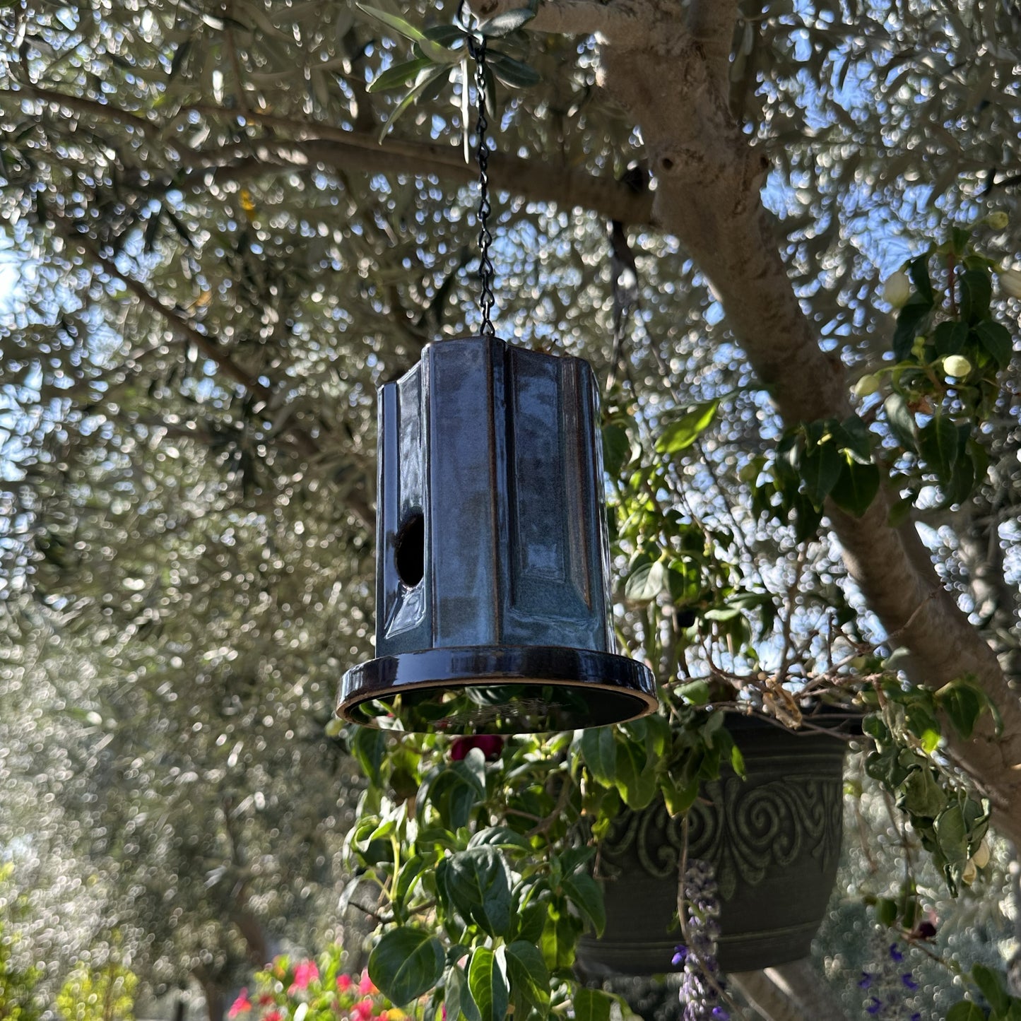 A hanging metal birdhouse with a weathered blue finish, suspended by a black chain in an outdoor garden setting with trees and plants in the background.