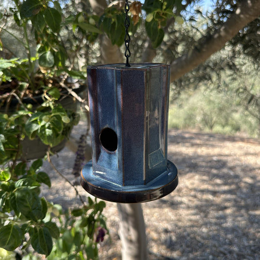 A hanging metal birdhouse with a weathered blue finish, suspended by a black chain in an outdoor garden setting with trees and plants in the background.