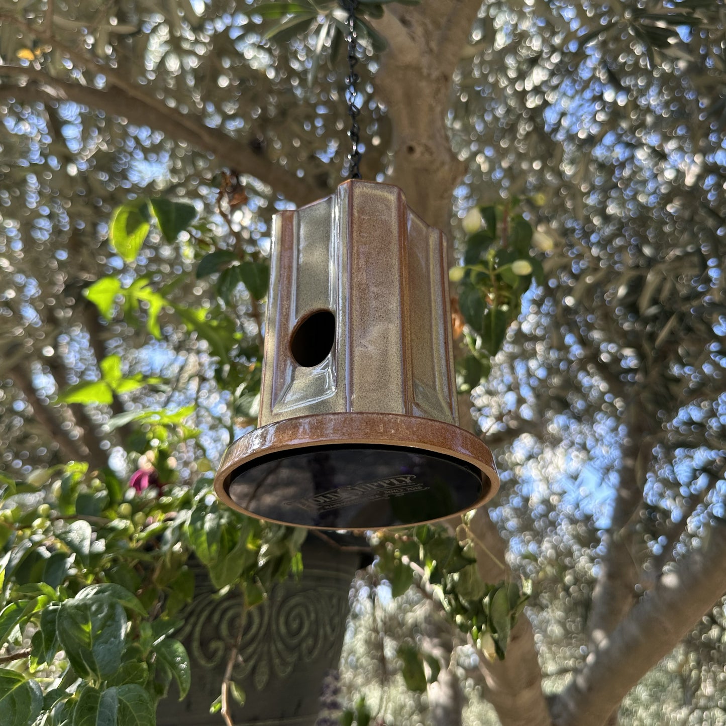 A hanging metal birdhouse with a weathered green finish, suspended by a black chain in an outdoor garden setting with trees and plants in the background.