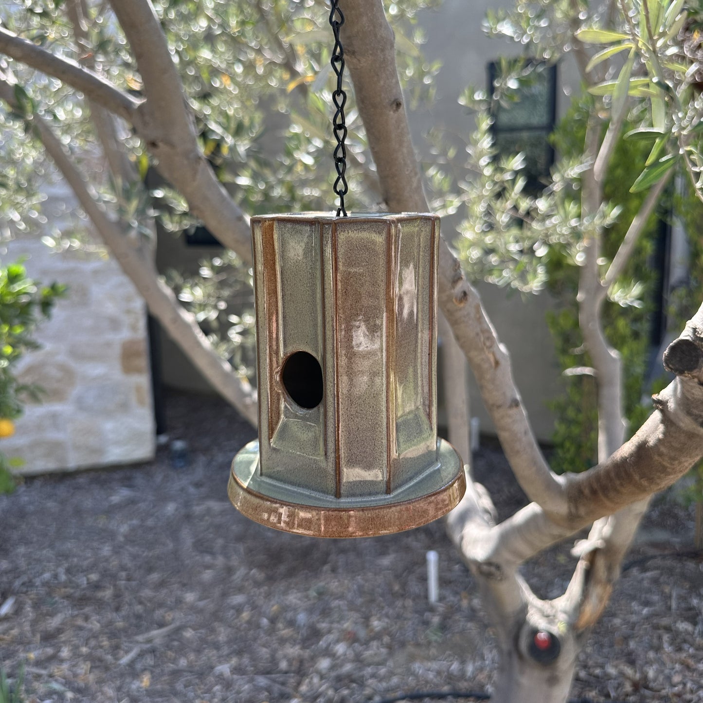 A hanging metal birdhouse with a weathered green finish, suspended by a black chain in an outdoor garden setting with trees and plants in the background.