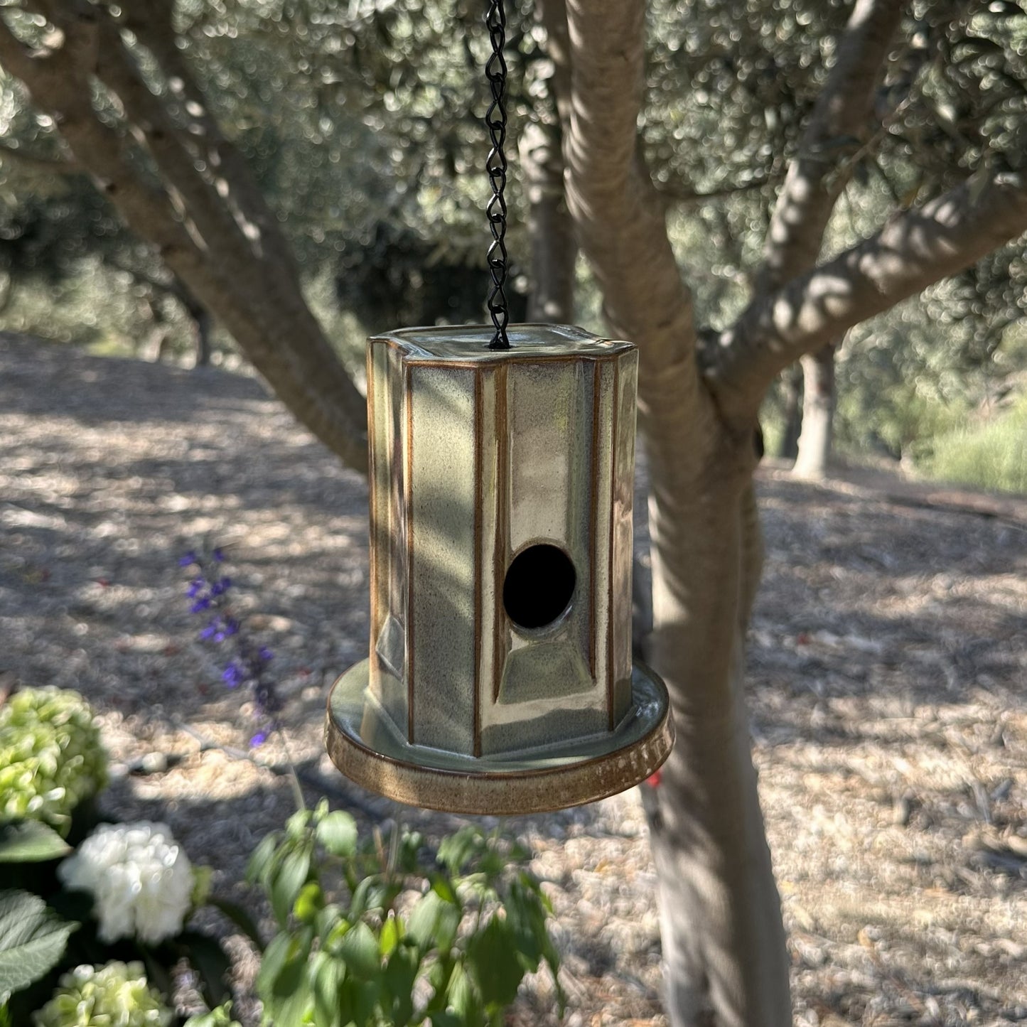 A hanging metal birdhouse with a weathered green finish, suspended by a black chain in an outdoor garden setting with trees and plants in the background.