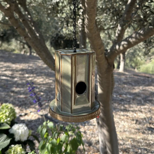 A hanging metal birdhouse with a weathered green finish, suspended by a black chain in an outdoor garden setting with trees and plants in the background.