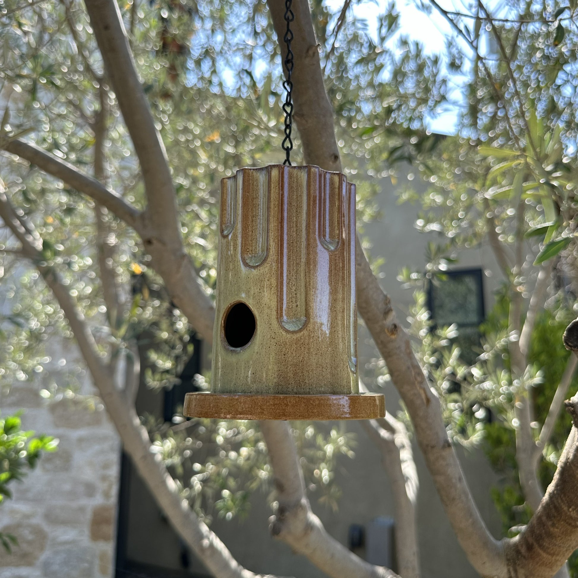 A hanging ceramic birdhouse with a glossy brown and beige drip-glaze finish, suspended by a black chain in an outdoor garden setting with olive trees and flowers in the background.