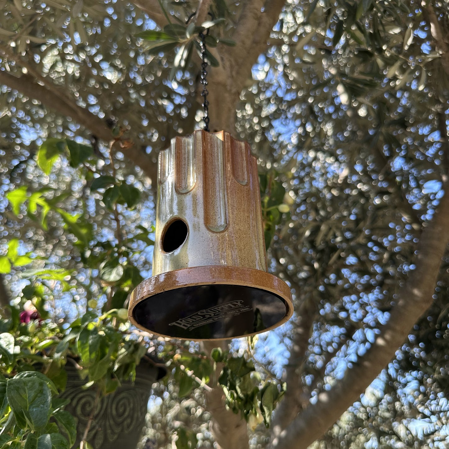 A hanging ceramic birdhouse with a glossy brown and beige drip-glaze finish, suspended by a black chain in an outdoor garden setting with olive trees and flowers in the background.
