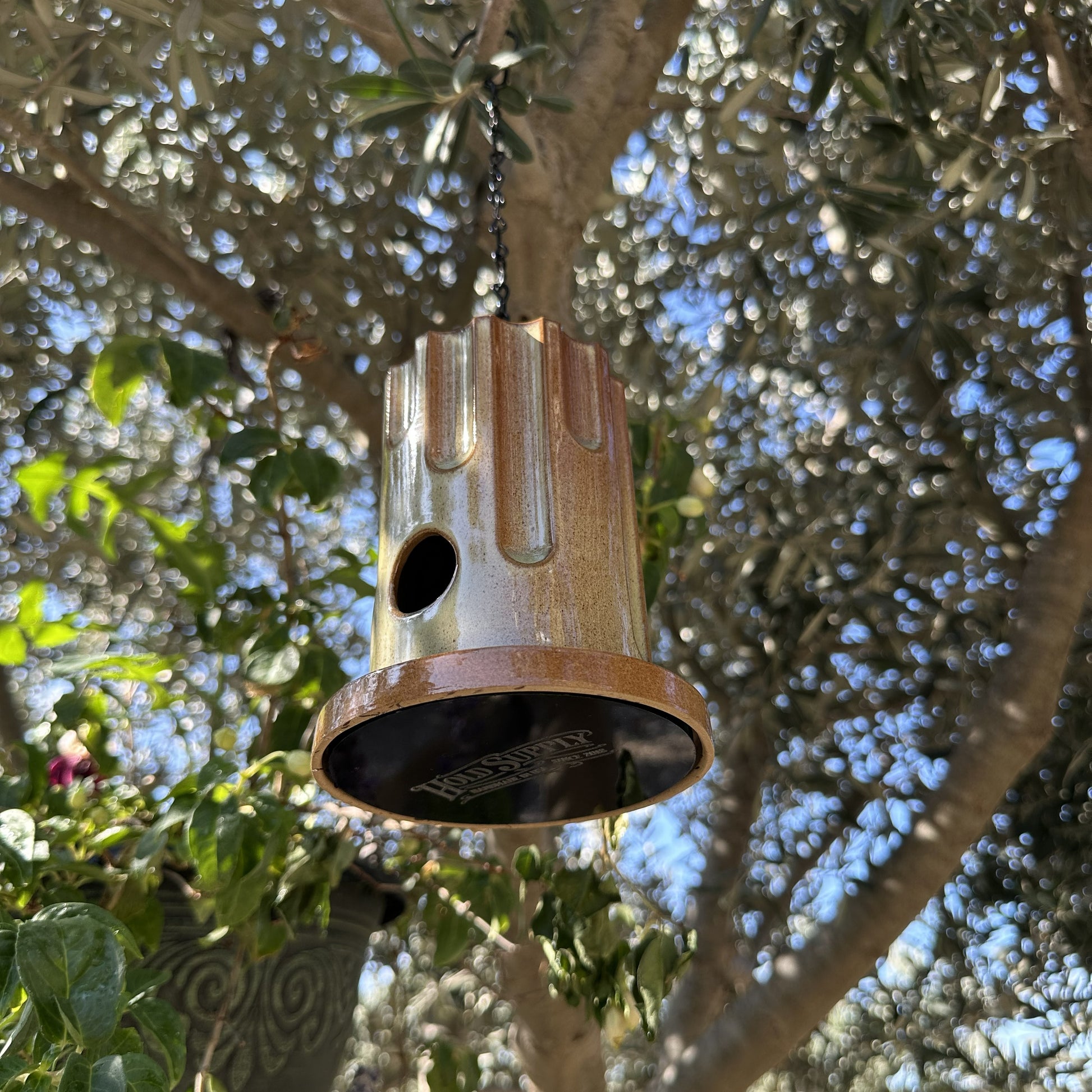 A hanging ceramic birdhouse with a glossy brown and beige drip-glaze finish, suspended by a black chain in an outdoor garden setting with olive trees and flowers in the background.
