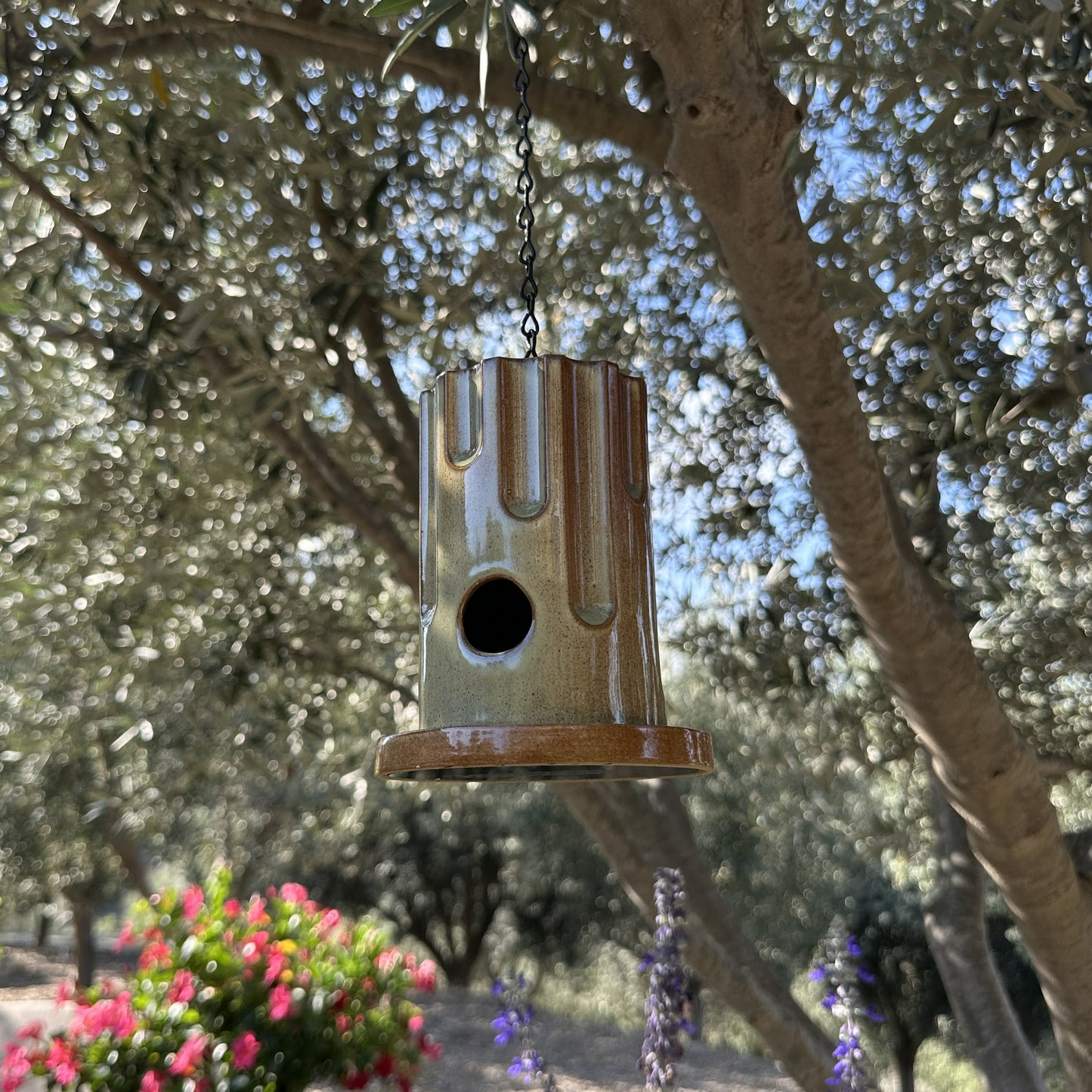 A hanging ceramic birdhouse with a glossy brown and beige drip-glaze finish, suspended by a black chain in an outdoor garden setting with olive trees and flowers in the background.