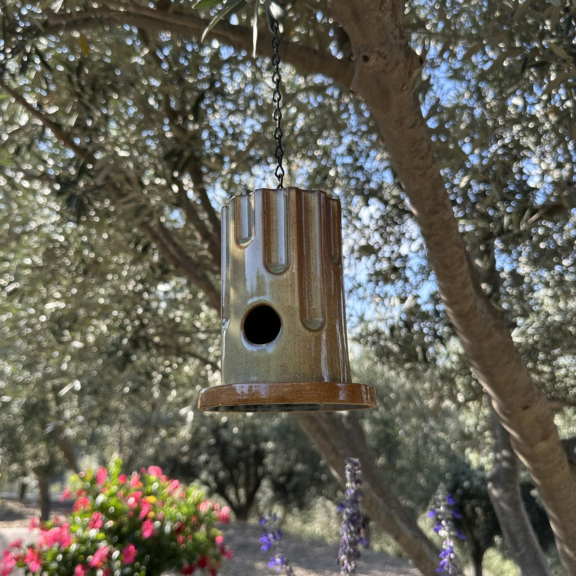 A hanging ceramic birdhouse with a glossy brown and beige drip-glaze finish, suspended by a black chain in an outdoor garden setting with olive trees and flowers in the background.