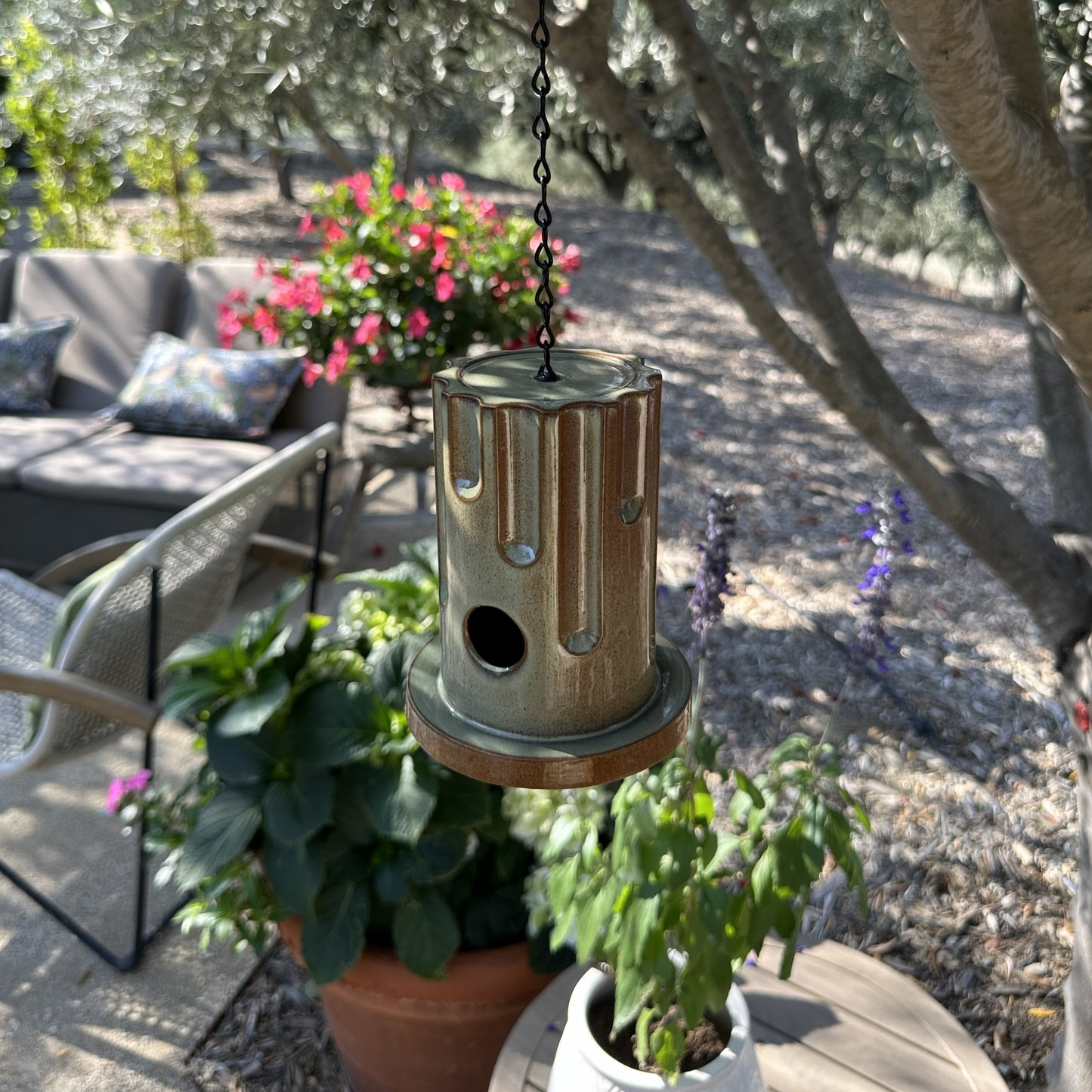 A hanging ceramic birdhouse with a glossy brown and beige drip-glaze finish, suspended by a black chain in an outdoor garden setting with olive trees and flowers in the background.