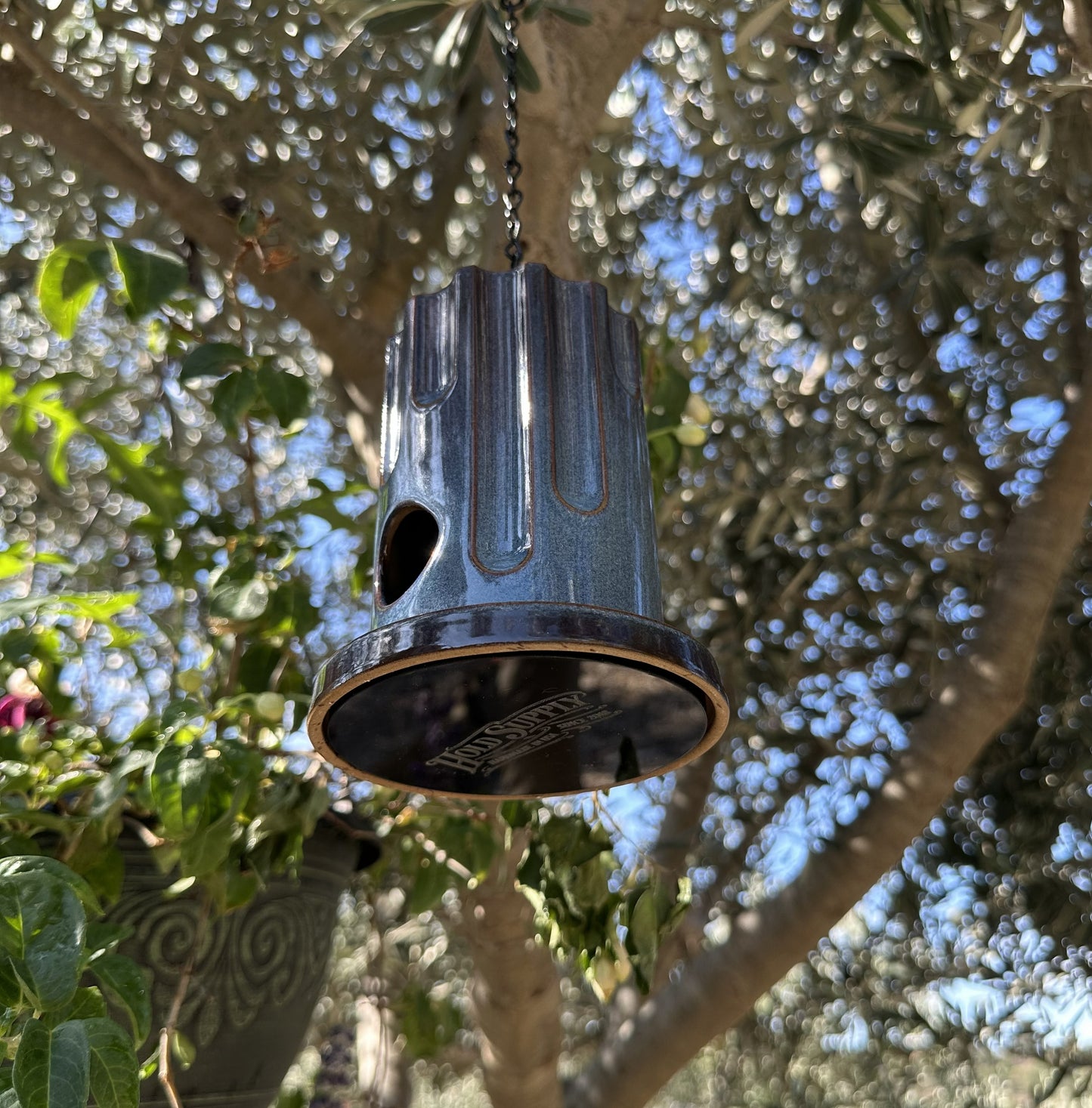 A hanging ceramic birdhouse with a glossy blue and red drip-glaze finish, suspended by a black chain in an outdoor garden setting with olive trees and flowers in the background.