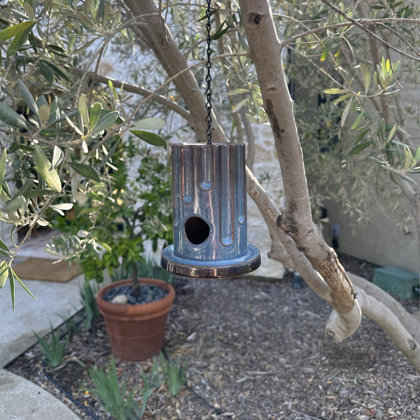 A hanging ceramic birdhouse with a glossy blue and red drip-glaze finish, suspended by a black chain in an outdoor garden setting with olive trees and flowers in the background.