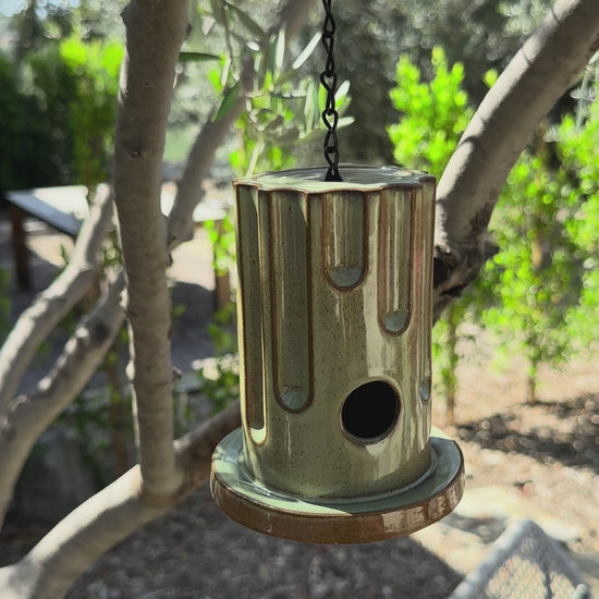A hanging ceramic birdhouse with a glossy brown and beige drip-glaze finish, suspended by a black chain in an outdoor garden setting with olive trees and flowers in the background.