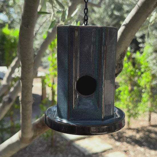 A hanging metal birdhouse with a weathered blue finish, suspended by a black chain in an outdoor garden setting with trees and plants in the background.