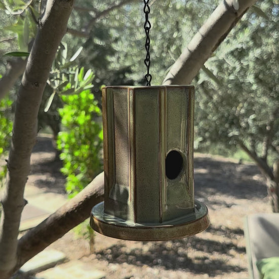 A hanging metal birdhouse with a weathered green finish, suspended by a black chain in an outdoor garden setting with trees and plants in the background.