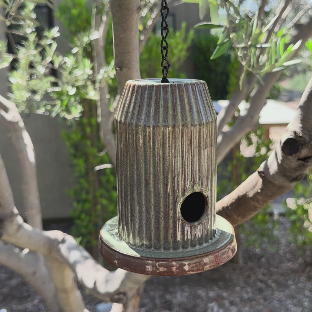 Green fluted ceramic birdhouse handcrafted from stoneware with black acrylic base, hanging outdoors.