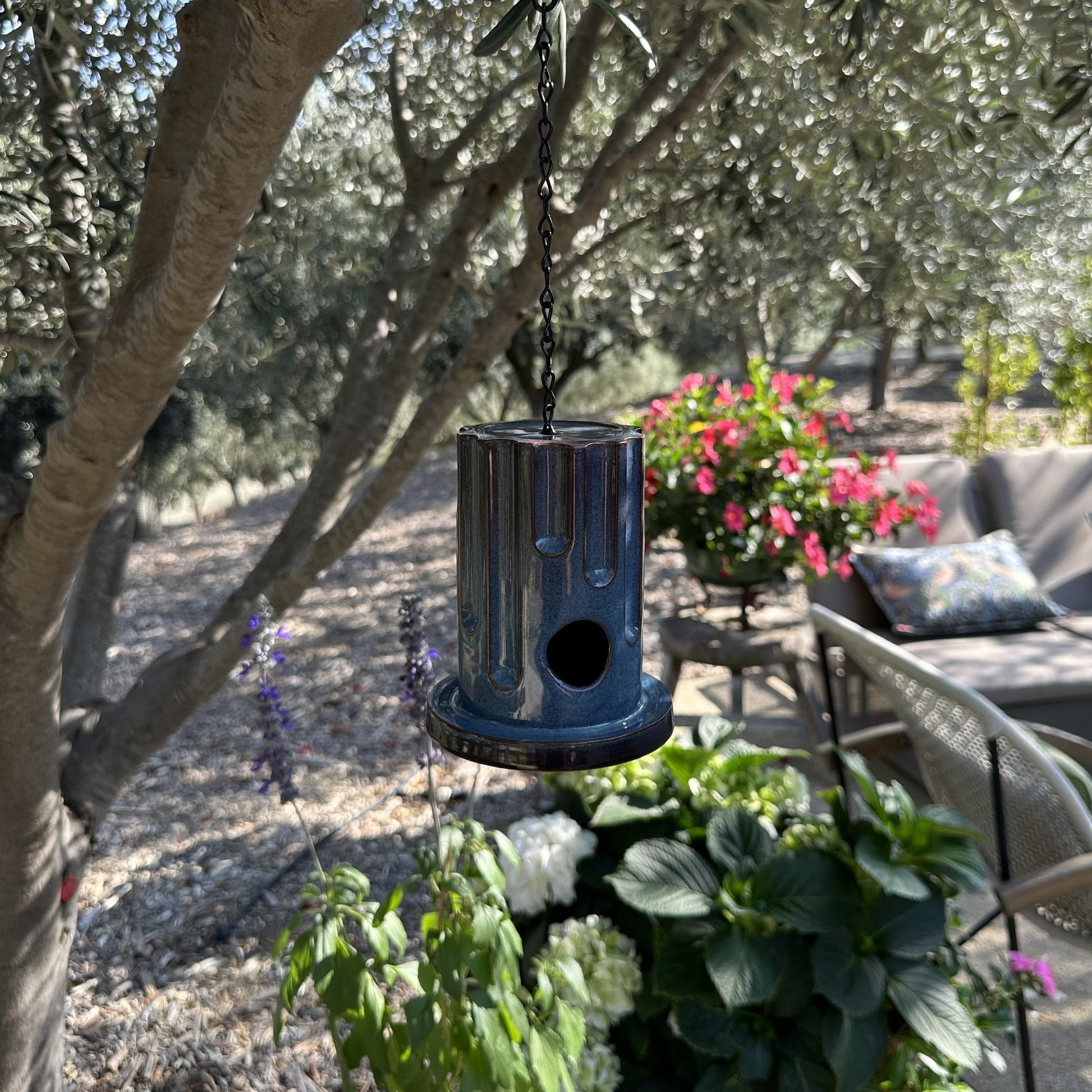 A hanging ceramic birdhouse with a glossy blue and red drip-glaze finish, suspended by a black chain in an outdoor garden setting with olive trees and flowers in the background.